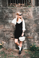 Portrait of a young blonde woman in schoolgirl clothes. Girl in a dress with sunglasses stands by an old wall in the park.
