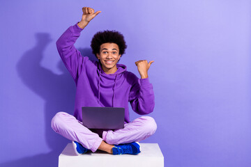 Full size photo of cheerful clever person sit on cube with laptop directing at offer empty space isolated on purple color background