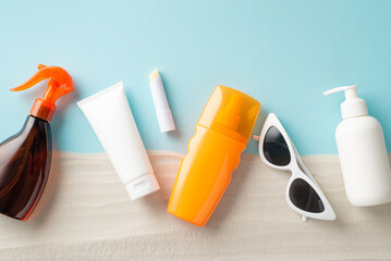 Top view composition comprising of sunblock oil, cream bottles, lip balm with SPF devoid of labels, along with sunglasses on pastel blue and sandy backdrop, featuring space for text or advertisement