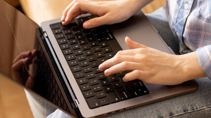 A teenage girl in casual clothes and blue jeans is typing on a laptop keyboard. A slender young freelance woman is sitting on the couch with a computer on her lap. Work at home. Close up.