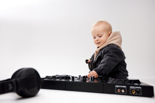 A cute kid in a beige hoodie and a funny black motorcycle jacket sits on the floor with dj headphones and a dj mixing console. Music and fun. Isolate on a white background.