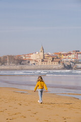 child girl walking on the beach with the town of Gijon, Asturias, in the background. She is wearing winter clothes, jumper and jeans. 