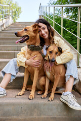 Charming young smiling girl is sitting on the steps in the park with two golden dogs on a sunny...