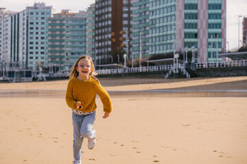 child girl running on the beach with the town of Gijon, Asturias, in the background. She is wearing winter clothes, jumper and jeans. 