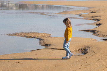 Kid blondie girl walking at the sand beach looking at the sea. She is wearing winter clothes, jumper and jeans.