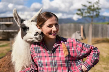 Rolgordijnen Lama Young woman in shirt standing next to white llama at zoo on a sunny day, smiling, posing for picture