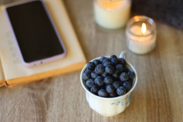 Cup of blueberries, set of books, phone, eyeglasses, lit candles and blanket on the table. Hygge at home. Selective focus.