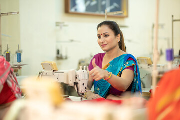 Indian woman showing thumbs up while work on sewing machine at textile factory.