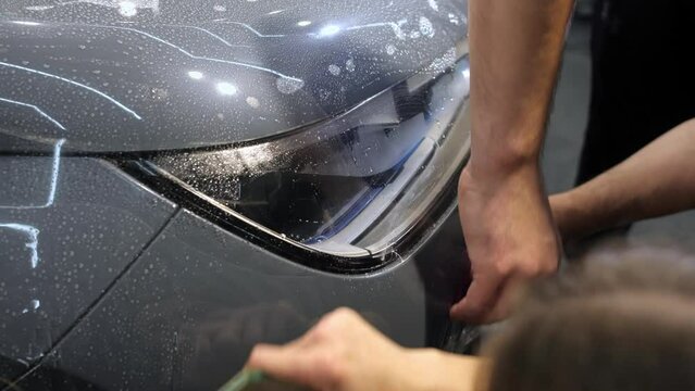 Female and male hands with plastic scrapers stick protective film on the bumper and headlight of a gray new car. Close up. Car body protection concept.