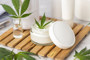 Obraz na płótnie Canvas Opened cream jar with white lid near green cannabis leaves close up, CBD cosmetic mockup