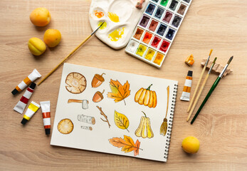 Photo of autumn watercolor drawing. Against the backdrop of a palette of brushes and tubes of paint on a light wooden background. Drawing of leaves and pumpkins, in warm processing