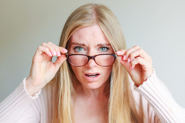 Shocked angry young woman in disbelief lowering glasses looking at camera with indignation isolated on grey blank studio background