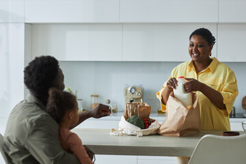 Happy African American mom taking out groceries from bags and talking to her family in the kitchen