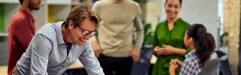 Happy businessman standing in the modern coworking space, looking at laptop screen and smiling while his young multiracial colleagues communicating on the background