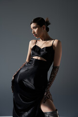 Fototapeta premium fashionable spring concept, young asian woman in black strap dress and animal print gloves posing with hand on hip and looking away on dark grey background