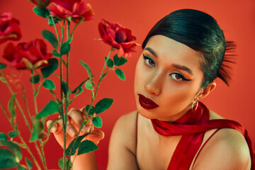 Fototapeta premium spring style photography, charming asian woman with brunette hair and bold makeup touching green leaves on roses and looking at camera on red background, generation z