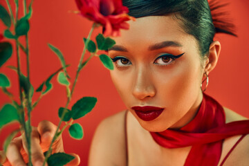 portrait of young asian woman in neckerchief, with bold makeup and expressive gaze looking at camera near flowers on red background, spring fashion photography, generation z