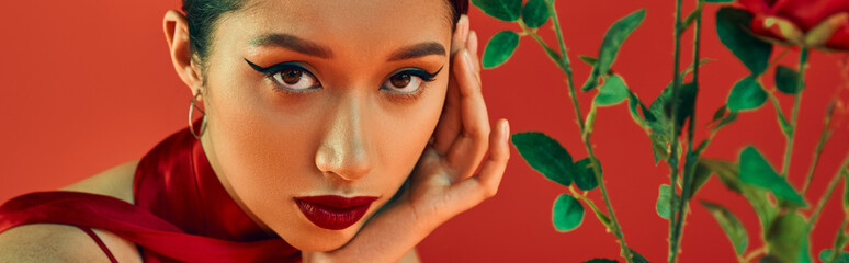 portrait of youthful and alluring asian woman with bold makeup and expressive gaze holding hand near face and looking at camera near flowers on red background, trendy spring, generation z, banner