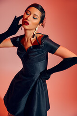 Fototapeta premium mesmerizing asian woman with bold makeup, brunette hair, in long gloves and black cocktail dress standing in stylish pose with closed eyes on red and pink background, spring fashion concept
