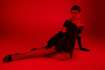 full length of young asian woman in stylish spring outfit sitting and looking away on vibrant background with red lighting effect, black cocktail dress, long gloves, fishnet tights, gen z fashion