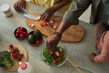 Top view of woman cutting vegetables on board for salad with her husband helping her