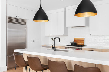 A kitchen detail with white and white oak cabinets, leather chairs sitting at an island, and a black and gold light hanging above. No names or brands.