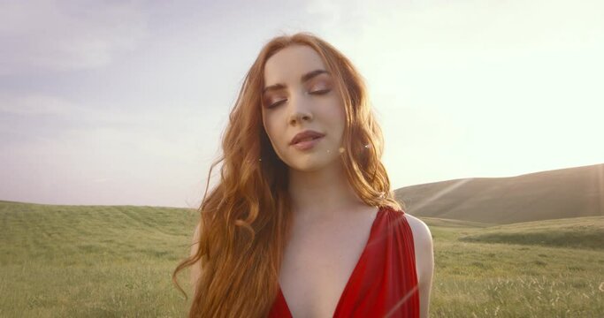 Slow motion of coquettish flirty red-haired woman in field waving fabric of red luxury fashion dress in front of camera lens. Woman flirtatiously waves red dress in front of camera at sunset in field.