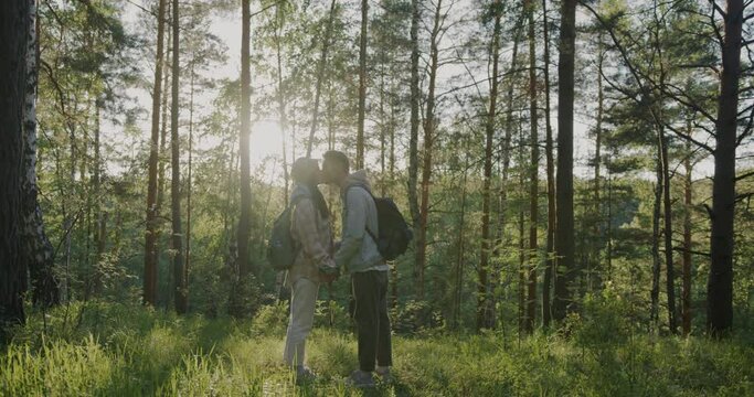 Slow motion of beautiful young couple kissing holding hands standing in sunlit forest enjoying nature. Loving relationship and tourism concept.