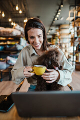 Beautiful and happy young woman sitting in modern pet shop cafe bar and enjoying in fresh coffee together with her adorable brown toy poodle.