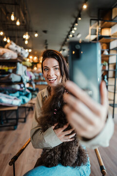 Beautiful and happy young woman sitting in modern pet shop cafe bar and enjoying in fresh coffee together with her adorable brown toy poodle. She also taking self portrait photo with smart phone.