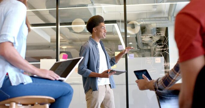 Happy biracial casual businessman presenting to diverse colleagues at glass wall, slow motion