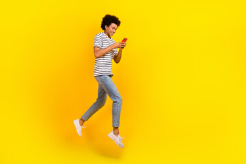 Fototapeta na wymiar Full body cadre of young addicted blogger chevelure guy jumping hold smartphone steps chatting online isolated on yellow color background