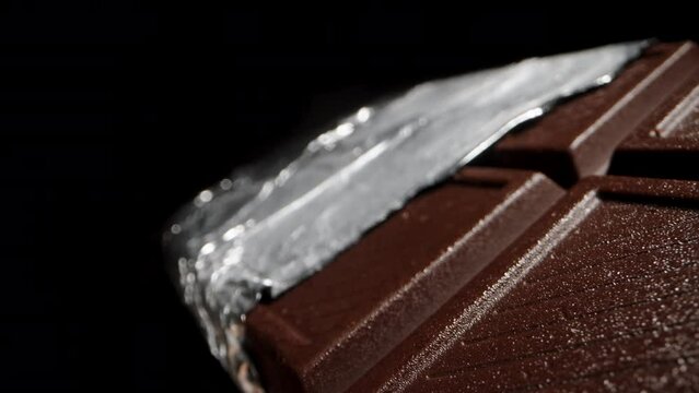 A large black chocolate bar, partially unwrapped from the foil, isolated on a black background. Dolly slider extreme close-up.