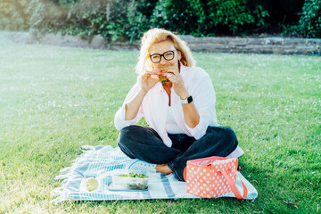 Middle aged caucasian woman sitting on green lawn and eating healthy lunch. Having picnic during...