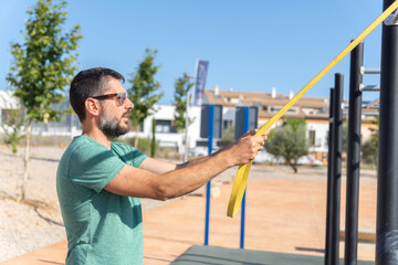 bearded man with sunglasses seen in profile training his arms with an elastic fitness band in an outdoor gym.