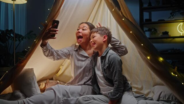 Little boy and his loving mom making faces taking self portrait on smartphone, sitting inside a tent