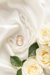 Two gold engagement rings on a satin ivory background with pearl beads and white roses. Vertical...