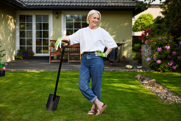 Happy mid adult woman standing with shovel in garden backyard looking around