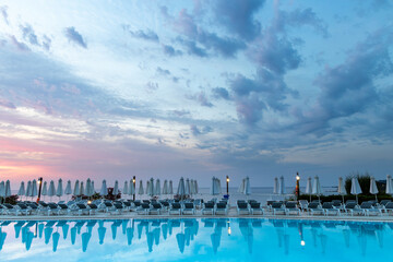 Fototapeta na wymiar Aegean sea sunrise behind pool, umbrellas and sun deck chairs at Kallithea, Greece