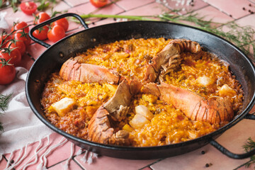 spanish seafood paella with prawns and squid pieces, traditional dish with rice in a hot pot, surrounded by fresh ingredients on a pink background table