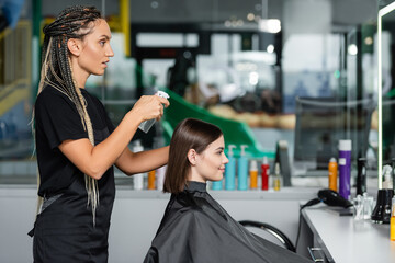 hairstylist spraying hair of female client, hairdresser with braids holding spray bottle near woman with short brunette hair in salon, hair cut, hair treatment, hair make over, hairdo, side view