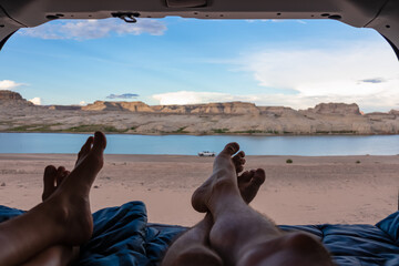 Leg of couple lying in camper van with panoramic sunset view of Wahweap Bay at Lake Powell in Glen Canyon Recreation Area, Page, Utah, USA. Sand beach on wild campground. Road trip romantic atmosphere