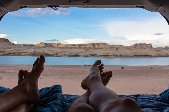 Leg Of Couple Lying In Camper Van With Panoramic Sunset View Of Wahweap Bay At Lake Powell In Glen Canyon Recreation Area, Page, Utah, USA. Sand Beach On Wild Campground. Road Trip Romantic Atmosphere