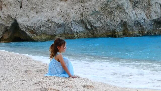 Little girl in summer blue dress watching at waves sitting on white sand on a beach with aquamarine sea. High quality FullHD footage