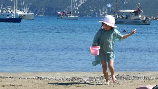 Little girl with summer hat playing with white sand and water building castle in a beach with aquamarine sea behind. High quality FullHD footage