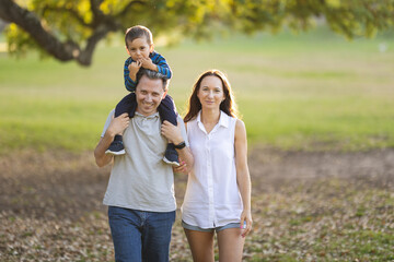 Fototapeta premium Cute white smiling family in the park - little son on the shoulders of his father and his wife standing by - looking in the camera
