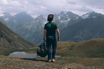 Fototapeta premium A man traveler with backpack and leather hat near the Koruldi Lake, Mestia in the Greater Caucasus Mountain Range Upper Svaneti, Georgia. Wildlife, travel and vacation. Summer day. Rear view