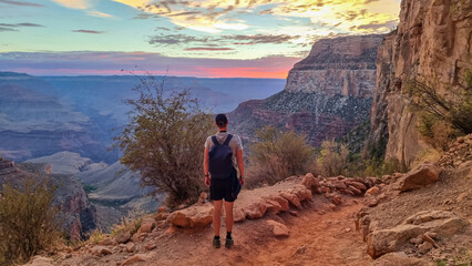 Rear view of man with backpack hiking along Bright Angel trail with panoramic aerial overlook of...