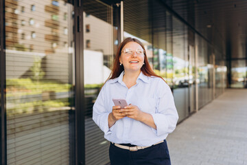 Fototapeta na wymiar Businesswoman using phone near office. Happy woman in a blue shirt walks on the street with her smartphone. Professional manager working on mobile device. Successful female using modern technology