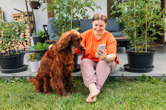 Beautiful Irish Setter Dog Is Laying On Grass. Cheerful Girl Having Video-call With Lover, Woman Holding Smartphone In Hand Shooting Selfie On Front Camera In The Yard Of The House. Full Body